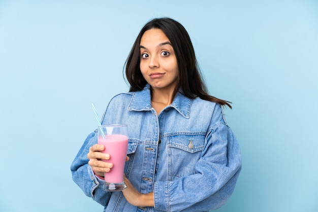 Mujer joven con batido de fresa sobre azul aislado haciendo gesto de dudas mientras levanta los hombros