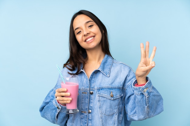 Mujer joven con batido de fresa sobre fondo azul aislado feliz y contando tres con los dedos