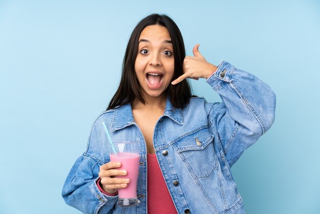 Mujer joven con batido de fresa sobre pared azul aislada haciendo gesto de teléfono. Llámame señal