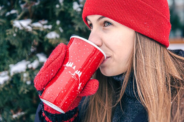 Una mujer joven bebe una bebida caliente de una taza térmica roja en invierno