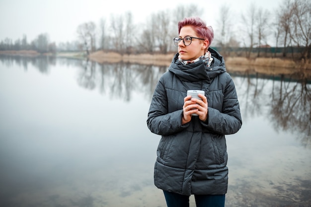 Mujer joven bebe taza de café en la orilla del lago