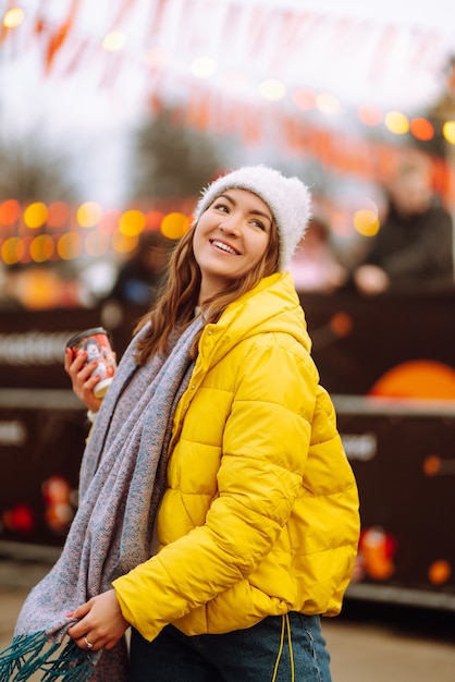 Mujer joven bebiendo café caliente mientras camina cerca del árbol de año nuevo.