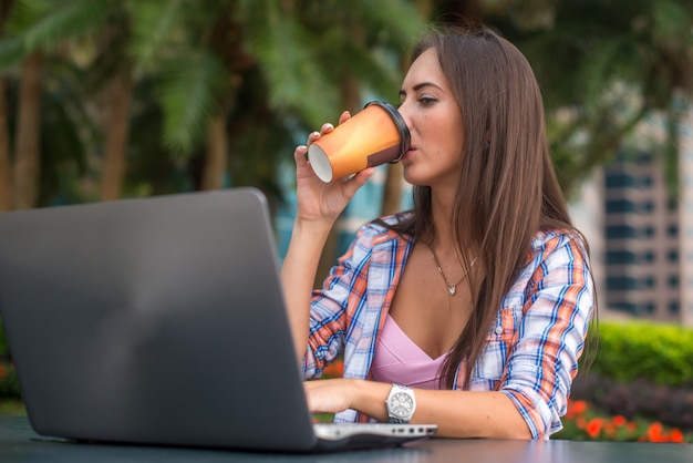 Mujer joven bebiendo café mirando la pantalla de su computadora portátil en el parque.