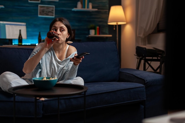 Mujer joven bebiendo cerveza de una botella y cambiando de canal de televisión para encontrar un programa o hacer una selección de películas. Adulto casual disfrutando de una bebida alcohólica y viendo un programa de televisión en el sofá.