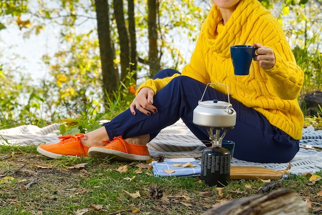 Mujer joven bebiendo su café recién hecho por la mañana afuera en el bosque sentado en la manta