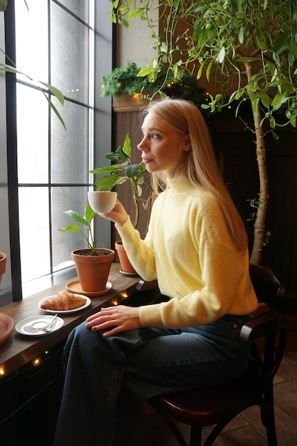 Mujer joven bebiendo té o café en una acogedora cafetería con plantas verdes