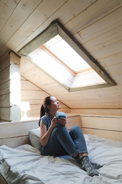 Mujer joven bebiendo té o café en la cama y mirando por la ventana por la mañana en casa