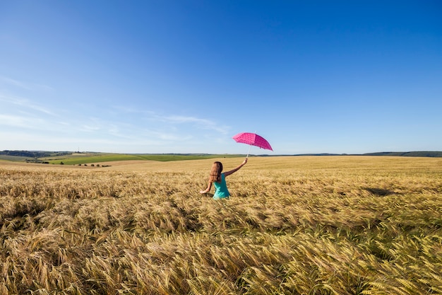 Mujer joven belleza al aire libre disfrutando de la naturaleza. Hermosa chica modelo adolescente en vestido azul con paraguas rojo en el campo de trigo en la luz del sol.