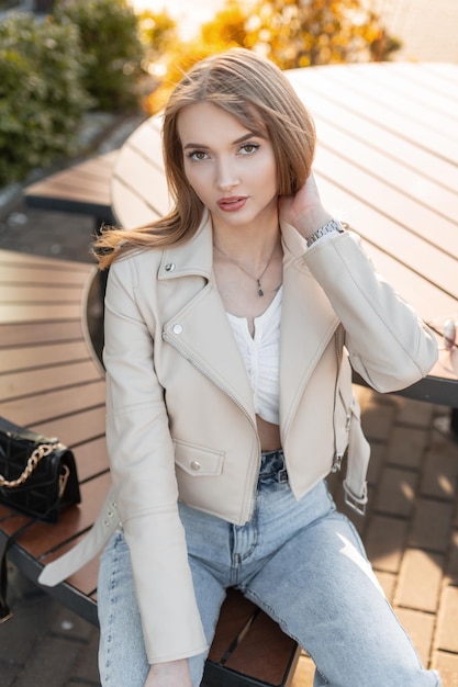 Mujer joven de belleza de moda con ropa de calle elegante con chaqueta de cuero de rock de moda y top con jeans vintage se sienta en el banco cerca de un café en la ciudad al atardecer