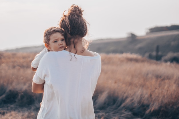 Mujer joven besa a su pequeño hijo al sol en el prado al atardecer. Joven madre sosteniendo a su bebé. Madre e hijo pequeño pasando un buen rato en la naturaleza. Imagen en tonos de color