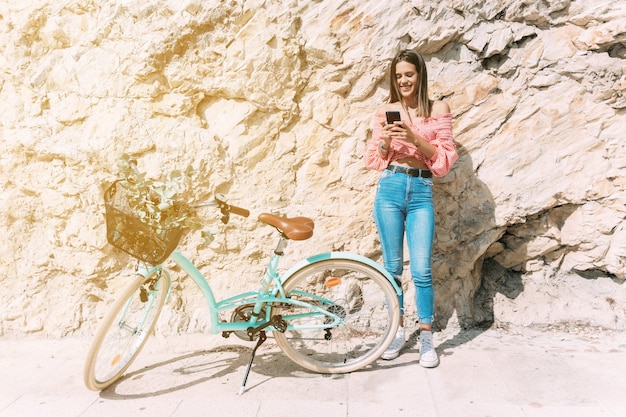 Mujer joven en una bicicleta charla con su teléfono móvil junto a un muro de piedra