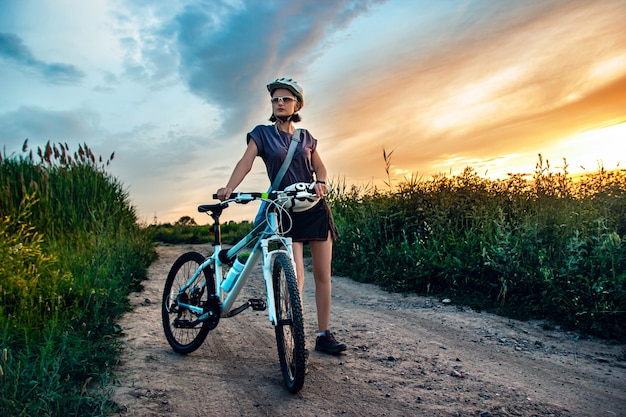 Mujer joven y bicicleta de montaña se encuentra en el campo verde rural en las noches de verano Aventura en bicicleta