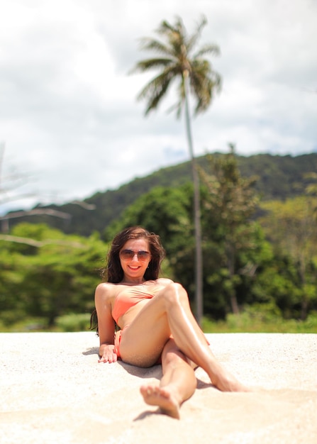 Mujer joven en bikini y gafas de sol, sentada en la arena de la playa, sonriendo, con las palmas detrás de ella.