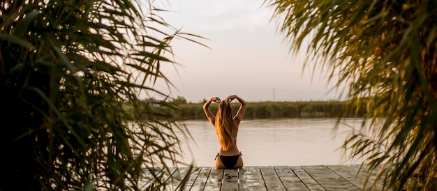 Mujer joven en bikini sentada en un muelle en el lago