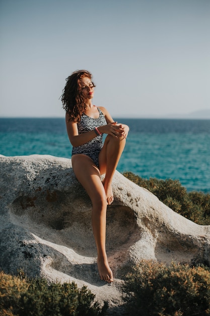 Mujer joven en bikini sentada en la roca junto al mar