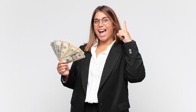 Mujer joven con billetes sintiéndose como un genio feliz y emocionado después de realizar una idea, levantando alegremente el dedo, ¡eureka!