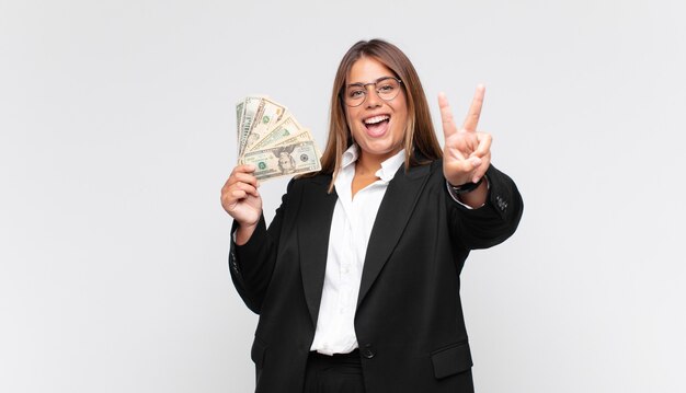 Mujer joven con billetes sonriendo y mirando feliz, despreocupada y positiva, gesticulando victoria o paz con una mano