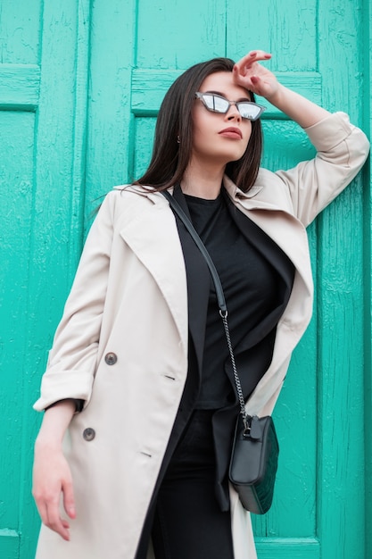 Mujer joven en blazer retro en camiseta negra de moda en gabardina de primavera de moda descansar cerca de la puerta de madera turquesa brillante vintage en la calle. Chica de moda en primer plano de ropa casual. Estilo juvenil.