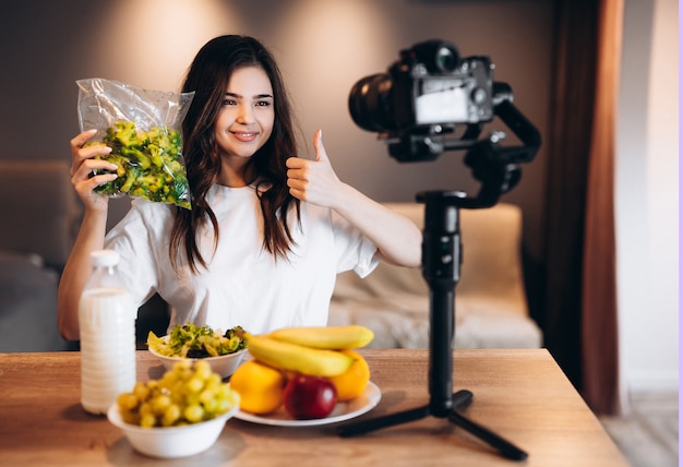 Mujer joven blogger de alimentos saludables cocinar ensalada vegana de frutas frescas en el estudio de la cocina, tutorial de filmación en la cámara para el canal de video. joven mujer influyente muestra su amor por la alimentación saludable.