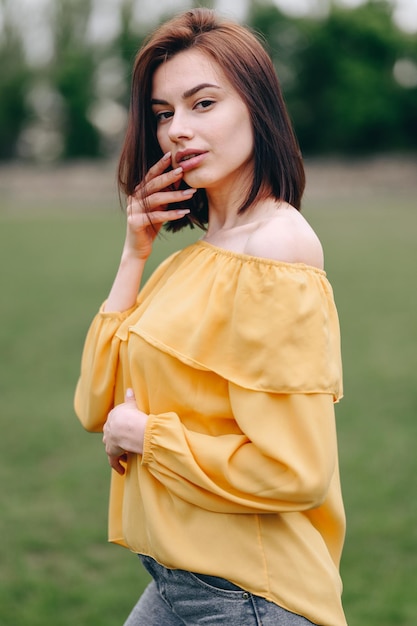 Una mujer joven con una blusa amarilla está de pie sobre un fondo natural Mano a los labios Retrato vertical