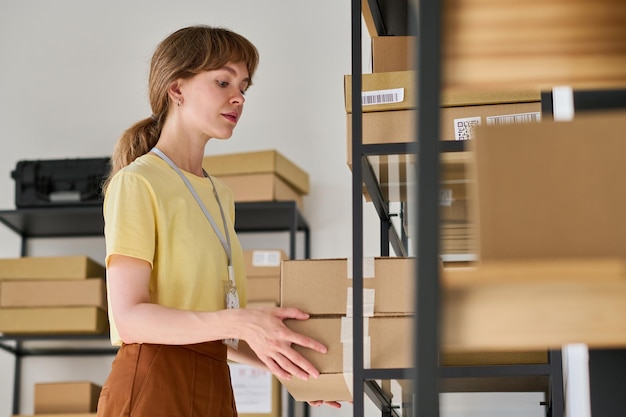 Mujer joven con blusa amarilla poniendo cajas en el estante del almacén mientras trabaja con pedidos empaquetados de clientes y clientes de tiendas en línea