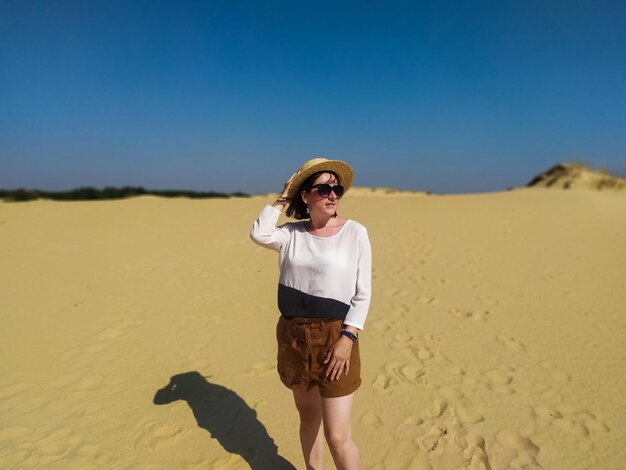 Mujer joven en blusa blanca y sombrero de paja posando en el desierto bajo el cielo azul Chica se encuentra en las dunas de arena en el desierto ucraniano Oleshky Sands parque natural Oleshkivski pisky Kherson oblast