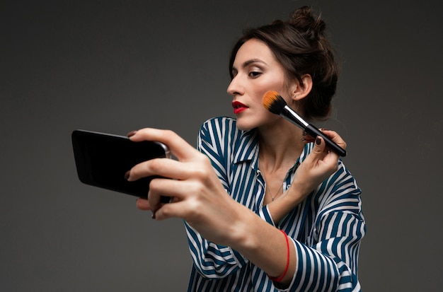 Mujer joven en blusa a rayas con teléfono celular negro en lugar de espejo y ponerse colorete con pincel de maquillaje aislado