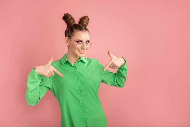 Mujer joven con bollos divertidos en camisa verde brillante apunta sus dedos índices hacia abajo Lugar para texto y publicidad