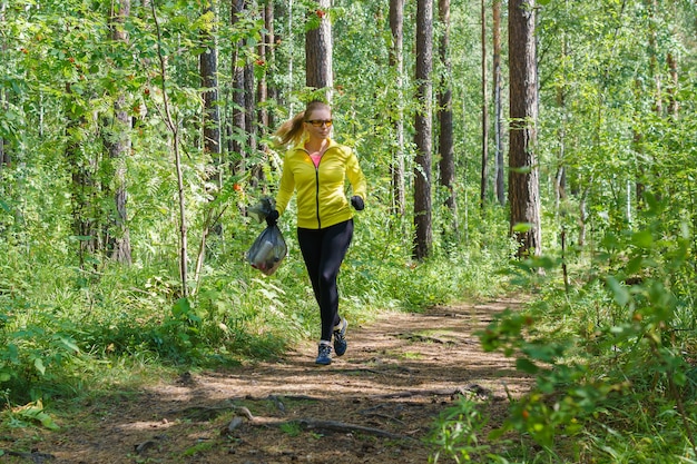 Mujer joven con una bolsa de basura en la mano trotar por un sendero forestal mientras ploging en un parque