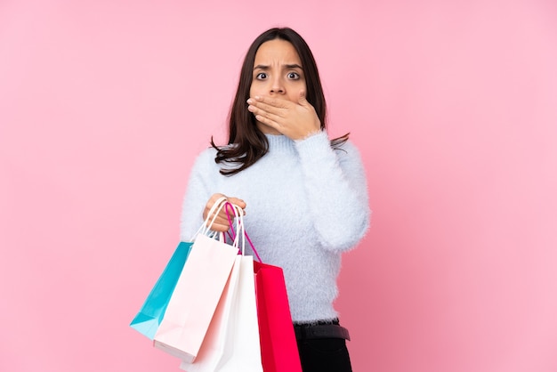 Mujer joven con bolsa de compras sobre pared rosa aislada que cubre la boca con las manos