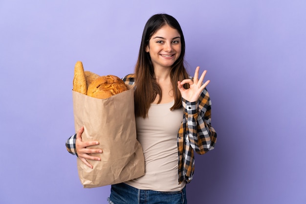 Mujer joven con una bolsa llena de panes aislados en púrpura mostrando un signo bien con los dedos