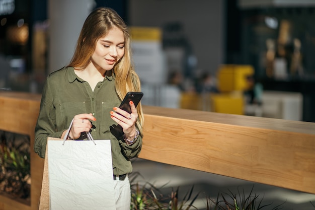 Mujer joven con bolsas de compras usando su teléfono inteligente para comprar