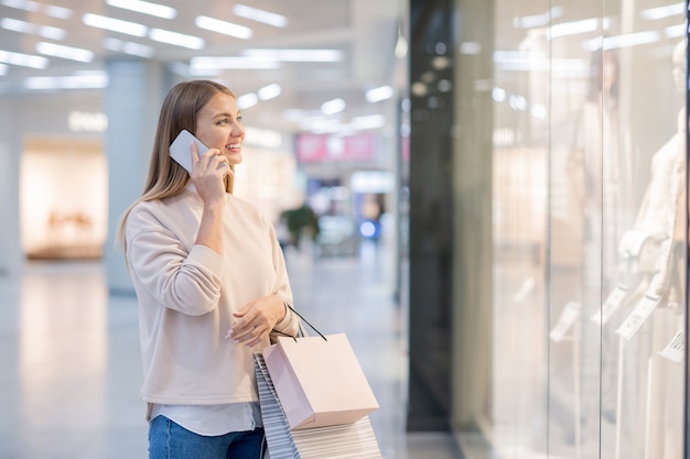 Mujer joven con bolsas de papel hablando con alguien por teléfono móvil mientras mira la nueva colección de moda en el escaparate