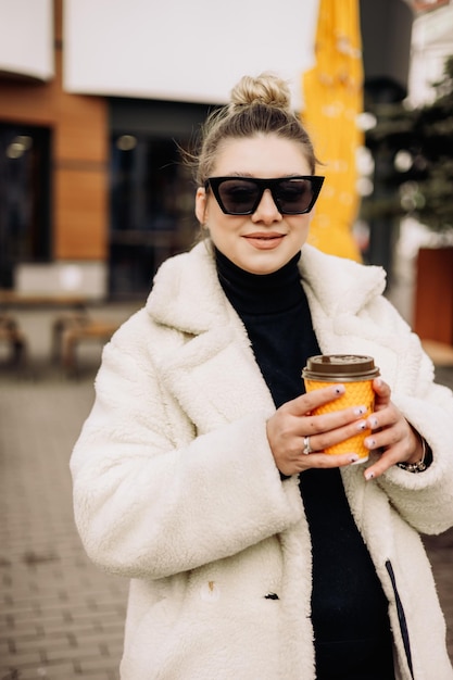 Una mujer joven y bonita con un abrigo de piel blanco y gafas de sol con café camina por la ciudad