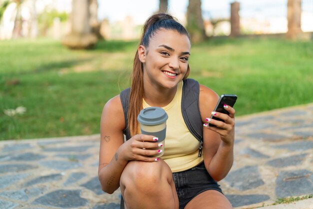 Mujer joven y bonita al aire libre usando teléfono móvil y sosteniendo un café para llevar