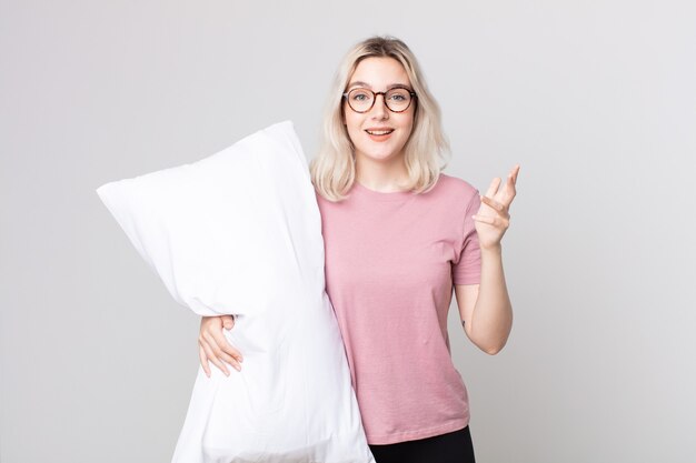 Mujer joven bonita albina que se siente feliz, sorprendida al darse cuenta de una solución o idea en pijama y sosteniendo una almohada