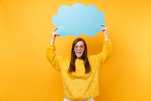 Mujer joven y bonita alegre en anteojos de corazón con azul en blanco vacío Diga nube, bocadillo de diálogo aislado sobre fondo amarillo brillante. Personas sinceras emociones, concepto de estilo de vida. Área de publicidad.