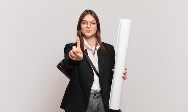 Mujer joven y bonita arquitecta sonriendo con orgullo y confianza haciendo la pose número uno triunfalmente, sintiéndose como un líder