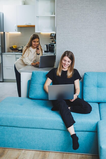 Una mujer joven y bonita está sentada en el sofá de casa y trabajando en una computadora portátil en el fondo. Otra mujer está hablando por teléfono en la cocina. Dos amigos trabajan en casa.