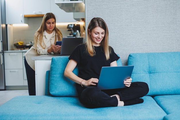 Una mujer joven y bonita está sentada en el sofá de casa y trabajando en una computadora portátil en el fondo. Otra mujer está hablando por teléfono en la cocina. Dos amigos trabajan en casa.