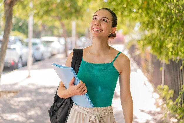 Mujer joven y bonita estudiante al aire libre mirando hacia arriba mientras sonríe