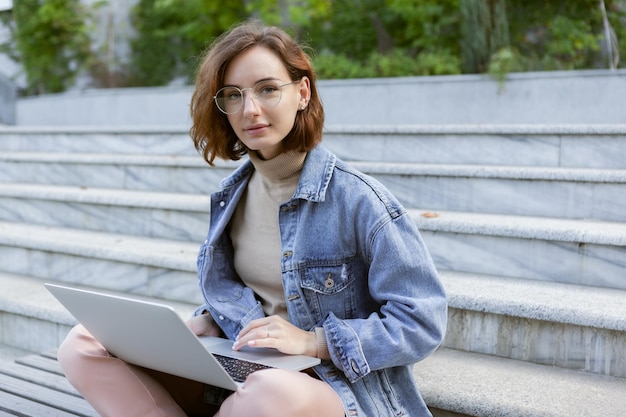 Mujer joven y bonita estudiante sentada con una laptop Chica caucásica trabajando o estudiando con una notebook al aire libre