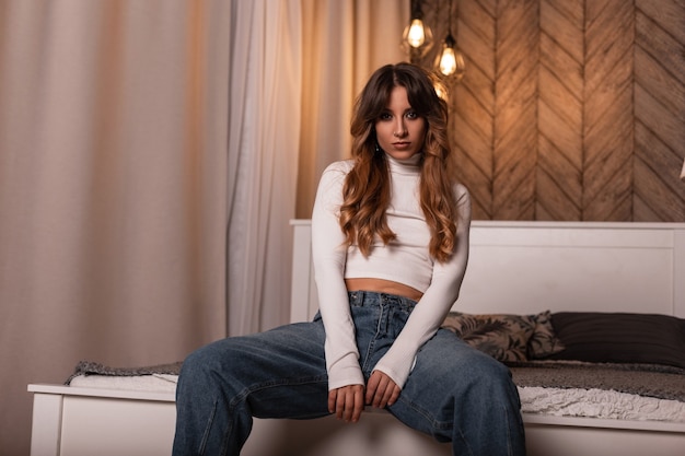 Mujer joven y bonita moderna con pelo largo rojo en ropa casual azul-blanca de moda juvenil posando sentada en una cama suave en el dormitorio. Modelo de moda chica sexy descansando en el interior.Belleza elegante dama en la habitación