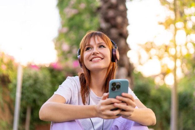 Mujer joven bonita pelirroja al aire libre escuchando música con el móvil y feliz