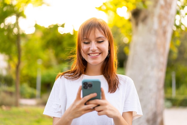 Mujer joven bonita pelirroja al aire libre usando teléfono móvil con expresión feliz