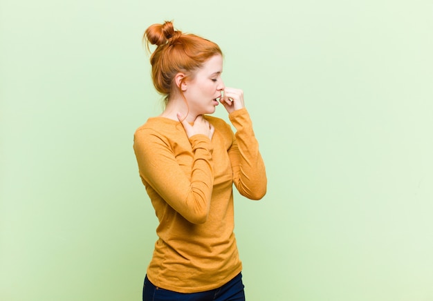 Mujer joven y bonita pelirroja que se siente enferma con dolor de garganta y síntomas de gripe, tos con la boca cubierta contra la pared verde