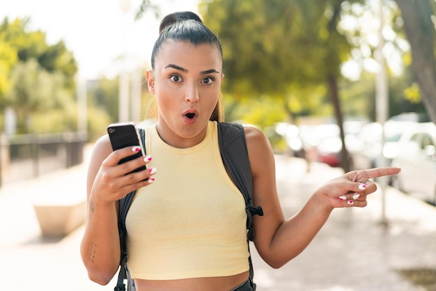 Mujer joven y bonita usando el teléfono móvil al aire libre sorprendida y apuntando hacia el lado