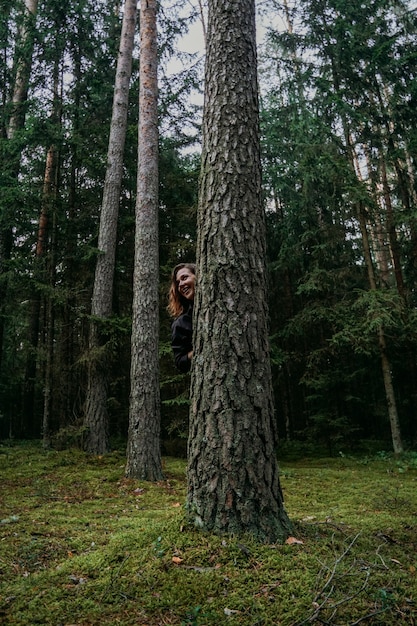 Una mujer joven en un bosque de coníferas mira desde detrás de un árbol