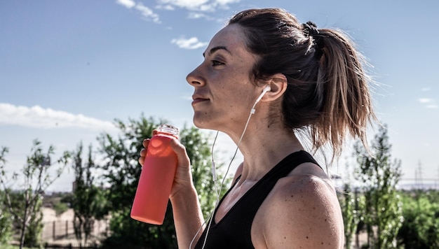 Mujer joven con botella de agua en la mano descansando después de correr mientras escucha música