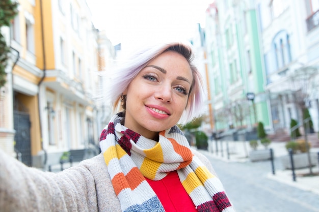 Mujer joven en una bufanda toma una selfie en el contexto de la ciudad